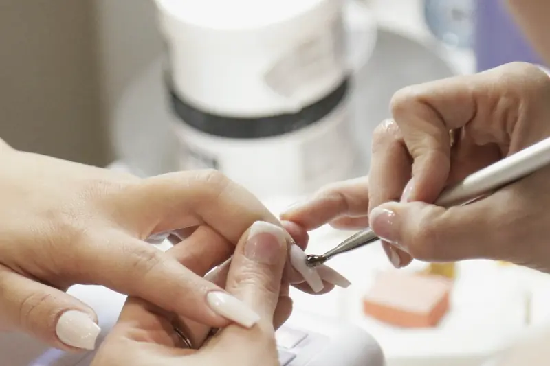 A nail technician pushes back a client’s cuticles with a metal tool, beside a white nail lamp.